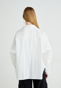 Persoon met lang blond haar, gekleed in een losse witte overhemd met knopen en zwarte broek, die met zijn rug naar de camera staat tegen een eenvoudige witte achtergrond.