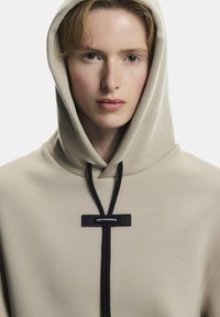 Beige kapucnis pulcsi fekete húzózsinórral, rajta egy téglalap alakú címke "SWISS ENGINEERING" felirattal. Puha tapintású, laza szabású és kapucnis kialakítású.