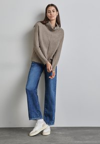 Beige Pullover mit Kapuze und strukturiertem Finish, kombiniert mit weit geschnittenen blauen Jeans und weißen Sneakers mit beigen Akzenten.