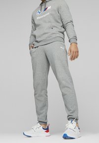 Grå sweatshirt med huva och BMW Motorsport-logotyp, tillsammans med matchande grå sweatshirtbyxor. Skorna har vit bas med röda och blå accenter.