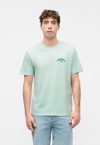 Νεαρός άντρας με σγουρά μαλλιά που φοράει ένα ανοιχτό πράσινο μπλουζάκι «Rip Curl» και ανοιχτό μπλε τζιν, στέκεται μπροστά σε λευκό φόντο.