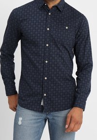 Marinblå skjorta med knappar, med diamantmönster, långa ärmar, krage och vita knappar. Bärs med ljusblå slitna jeans.