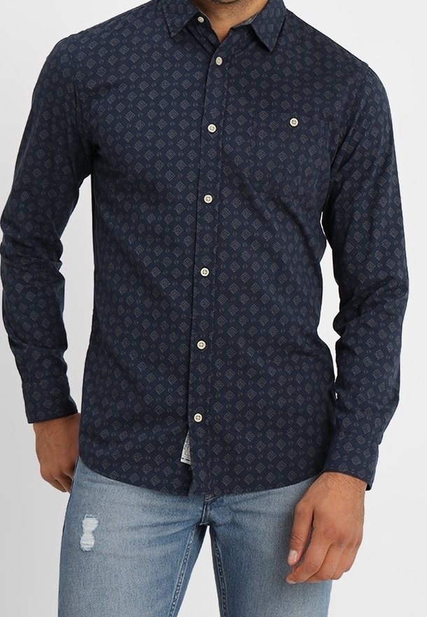 Marinblå skjorta med knappar, med diamantmönster, långa ärmar, krage och vita knappar. Bärs med ljusblå slitna jeans.