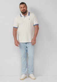 Kremowa koszulka polo z niebieskim kołnierzykiem i akcentami na rękawach, zestawiona z jasnoniebieskimi dżinsami i białymi trampkami. Casualowy fason, jednolity kolor, minimalne logo.