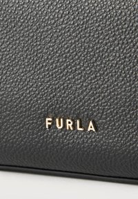 Μαύρη επιφάνεια από δερμάτινη υφή με μικρά χρυσά γράμματα που σχηματίζουν το όνομα της μάρκας «FURLA», τοποθετημένα στο κέντρο κοντά στο κάτω μέρος.