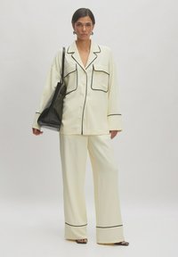 Crème-kleurige pyjama set met zwarte piping details, bestaande uit een loszittend overhemd met knopen en wijde broekspijpen. Gecombineerd met een zwarte tas.