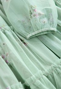 Tessuto verde chiaro e trasparente con ricami floreali rosa e sezioni arricciate elastiche, creando una delicata texture a strati.