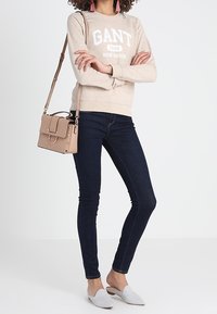 Sudadera beige con letras blancas, jeans oscuros ajustados, zapatos de lona gris claro y un pequeño bolso cruzado beige con herrajes dorados.