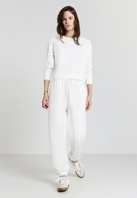 Donna con capelli lunghi che indossa una felpa bianca e pantaloni della tuta, che cammina in avanti su uno sfondo bianco semplice.