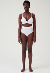 Witte triade bralette en hoogwarme ondergoedset met een gladde stof en eenvoudig ontwerp. Model staat blootsvoets tegen een neutrale achtergrond.