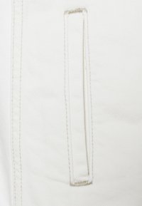 Vitt denimtyg med en sydd fickdetalj. Texturen är något ribbad och har kontrasterande beige sömmar längs fickkanterna.