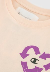 Perzikkleurig katoenen T-shirt met een paars, rond recyclen-logo en een zwart "C"-icoon in het midden, met een ribgebreide ronde hals.