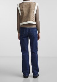 Person som bär en brun ribbad långärmad tröja, beige shearlingväst, mörkblå raka jeans och svarta chunky skor, stående med ryggen vänd.