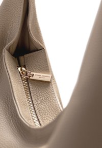 Innenansicht einer tanfarbigen Leder-Handtasche mit goldenem Reißverschluss und Markenanhänger. Texturierte Oberfläche und weiche Innenauskleidung sind sichtbar.