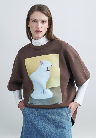 Bruine oversized sweatshirt met een kattenafbeelding op de voorkant, gecombineerd met een witte coltrui met lange mouwen en een denim rok.