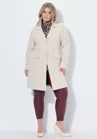 Una donna curvy che indossa un cappotto in velluto a coste beige, una maglietta con stampa leopardata, leggings in pelle bordeaux e scarpe con tacco bianche, in piedi contro un muro bianco.