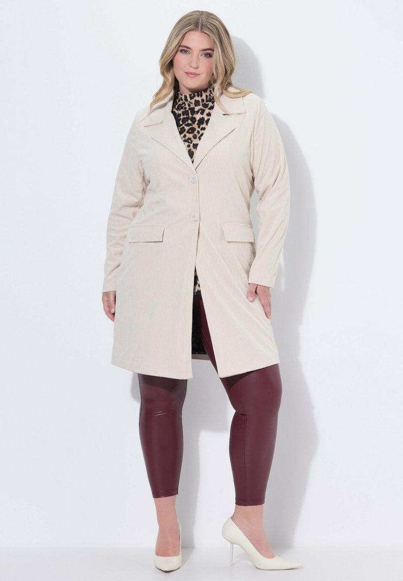 Una donna curvy che indossa un cappotto in velluto a coste beige, una maglietta con stampa leopardata, leggings in pelle bordeaux e scarpe con tacco bianche, in piedi contro un muro bianco.