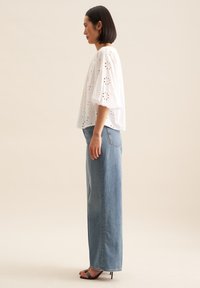Blusa bianca di cotone con ricamo ad occhiello e maniche a sbuffo, abbinata a jeans in denim azzurro chiaro a vita alta. Semplici sandali aperti.