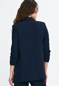 Granatowa marynarka wykonana z gładkiego materiału, charakteryzująca się dopasowanym krojem, podwiniętymi rękawami oraz pojedynczym rozcięciem z tyłu dla lepszej struktury.
