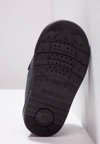 Semelle de chaussure en caoutchouc noir avec texture nervurée, dotée de trous de drainage et de texte en relief "GEOX" et "SOFTLY CUSHIONED 21." Design léger.