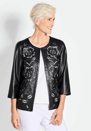 Veste en simili cuir noir avec des motifs floraux découpés sur le devant, encolure ronde et manches trois quarts, portée sur un haut blanc.