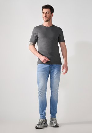 Šedé tričko s krátkým rukávem, modré slim-fit džíny, šedé a černé sportovní boty. Model stojí s jednou rukou ven, na světlém pozadí.