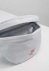 Sac en tissu gris avec une forme arrondie, une ouverture zippée, une texture lisse et un logo Helly Hansen rose et blanc bien visible sur le côté.