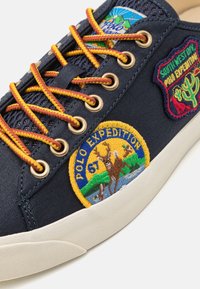 Baskets en toile bleu marine avec œillets dorés, lacets multicolores et patchs brodés illustrant des scènes de la nature et des textes. Semelle en caoutchouc blanche.