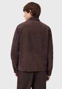 Brązowa dżinsowa kurtka z klasycznym kołnierzem, długimi rękawami i mankietami na guziki. Posiada lekko teksturowany materiał i detal w kształcie juka na plecach.