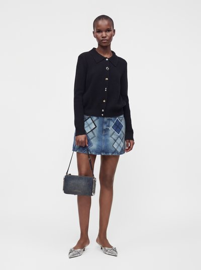 Femme portant un cardigan noir boutonné, une jupe en jean bleue avec patchwork, des ballerines argentées pointues et tenant un petit sac à main sombre.