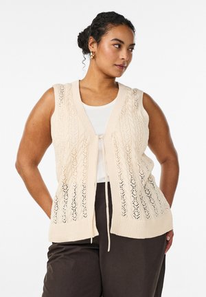 Žena s tmavými kučeravými vlasmi zviazanými do drdolu, s náušnicami zlatých kruhov, krémovou pletenou bezrukávovou vestou cez biele tričko a tmavohnedými nohavicami, hľadí doprava.