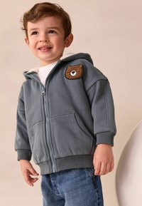 Småbarn som ler, iklädd en grå zip-up hoodie med ett teddy-björn märke, vit skjorta och blå jeans mot en beige bakgrund.