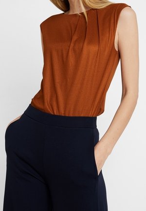 Ärmellose, rostrote Bluse, in eine hoch taillierte marineblaue Hose gesteckt, mit den Händen der Modellin in den Taschen, vor einem schlichten Hintergrund.