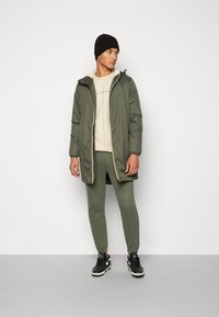 Olive green padded coat with a hood, zip closure, and contrast lining; beige shirt, olive joggers, and black sneakers. Plain background.
