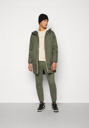 Olivengrøn polstret frakke med hætte, lynlås og kontrastfor; beige skjorte, olivengrønne joggers og sorte sneakers. Ensfarvet baggrund.