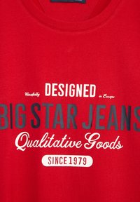 Raudona medvilninė marškinėlė su apvali apykakle, kuriame yra ryškus baltas ir juodas tekstas: "BIG STAR JEANS" ir "Kokybiškos prekės nuo 1979 metų."