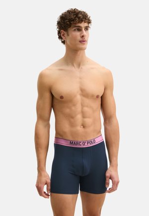 Männliches Model mit lockigem Haar, das marineblaue Boxershorts mit einem pinkfarbenen Bund trägt, auf dem "MARC O' POLO" steht, vor einem einfarbigen Hintergrund stehend.