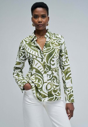 Camicia button-up con un motivo floreale verde e bianco, realizzata in tessuto morbido. Il modello indossa la camicia con pantaloni bianchi, con le mani nelle tasche.