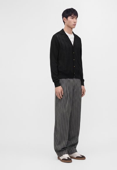 Giovane uomo in piedi, indossa un cardigan nero, camicia bianca, pantaloni larghi grigi a righe sottili e scarpe marroni e bianche su uno sfondo bianco semplice.