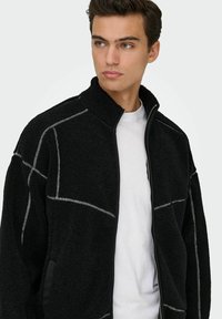 Veste en polaire noire avec un col haut, des coutures blanches contrastantes, fermeture zip et poches latérales. Texture douce et coupe décontractée.