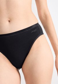 Nahaufnahme einer Person, die schwarze Sloggi-Unterwäsche trägt, mit Fokus auf der Taille und dem oberen Oberschenkel vor einem schlichten Hintergrund.