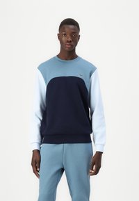 Sweter w kolorze granatowym i niebieskim z okrągłym dekoltem i długimi rękawami. Materiał jest miękki; na piersi znajduje się małe zielone logo.