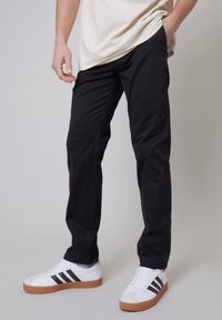 Pantaloni di cotone neri con una vestibilità dritta, abbinati a sneakers bianche con tre strisce marroni. Texture liscia, design minimale.