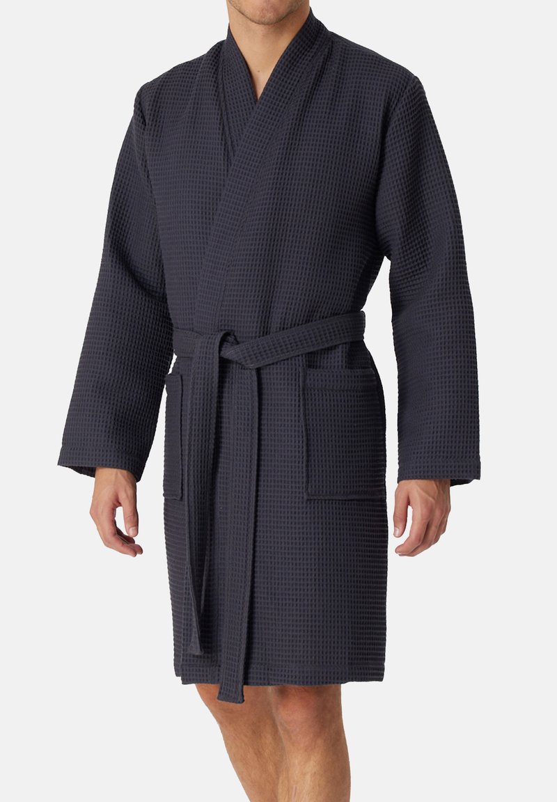 Mężczyzna ubrany w ciemnogranatowy szlafrok o fakturze wafla, z długimi rękawami, przednimi kieszeniami oraz przewiązanym paskiem, stojący na jednolitym tle.