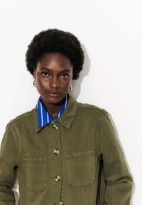 Jeune femme noire aux cheveux bouclés courts, portant des boucles d'oreilles en or, une chemise rayée bleue et une veste verte à boutons, sur fond blanc.