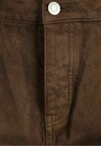 Los pantalones de mezclilla marrones presentan una superficie texturizada, cierre de botón plateado y detalles de costura a lo largo de los bolsillos y las costuras.