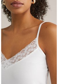 Balta kamizole ar V veida kakla izgriezumu un mežģīņu apdari. Audums ir gluds, ar plānām regulējamām siksnām un delikātām ziedu mežģīnēm.