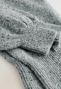 Manche de pull en tricot gris repliée sur le corps assorti, montrant un poignet côtelé et un fil texturé avec de petites speckles noires et bleues.