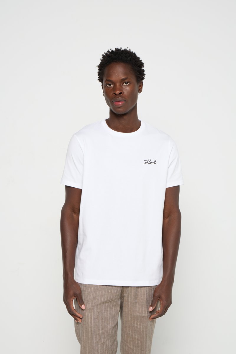 Camiseta blanca de algodón con cuello redondo, manga corta y logo negro bordado en el lado izquierdo del pecho. Llevada con pantalones de rayas finas en color marrón claro.