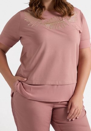 Mujer con camiseta de manga corta color rosa polvoriento con bordado de hoja de palma dorada y pantalones a juego, mano sosteniendo el dobladillo de la camiseta.